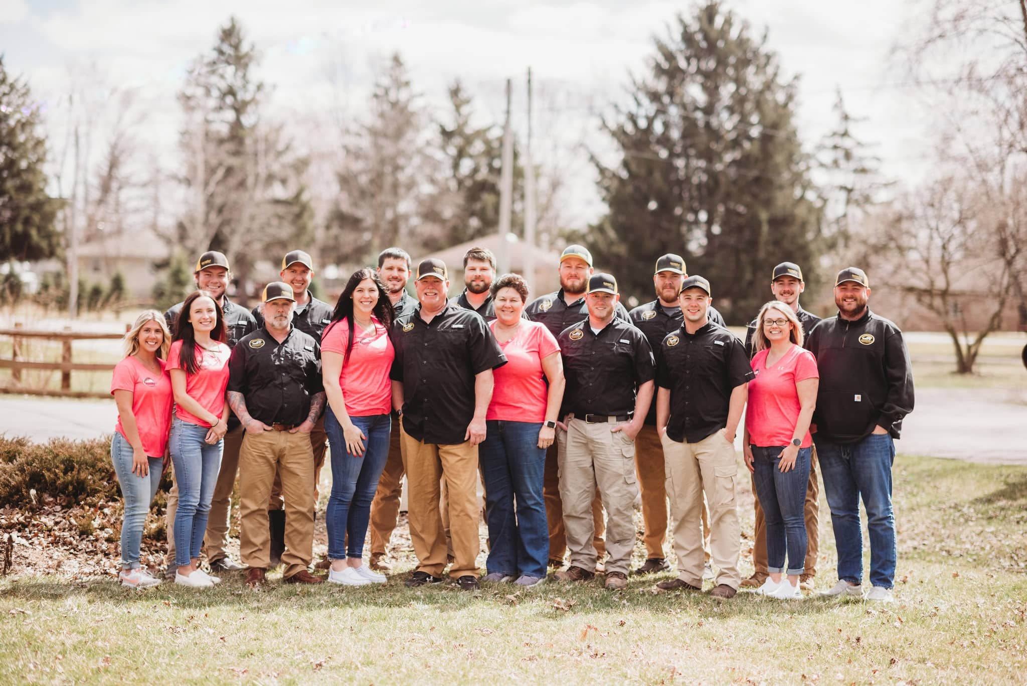 Group portrait of a InsideOut Inspections Plus team posing outdoors on a grassy lawn in matching black and pink shirts with a backdrop of tall pine trees.