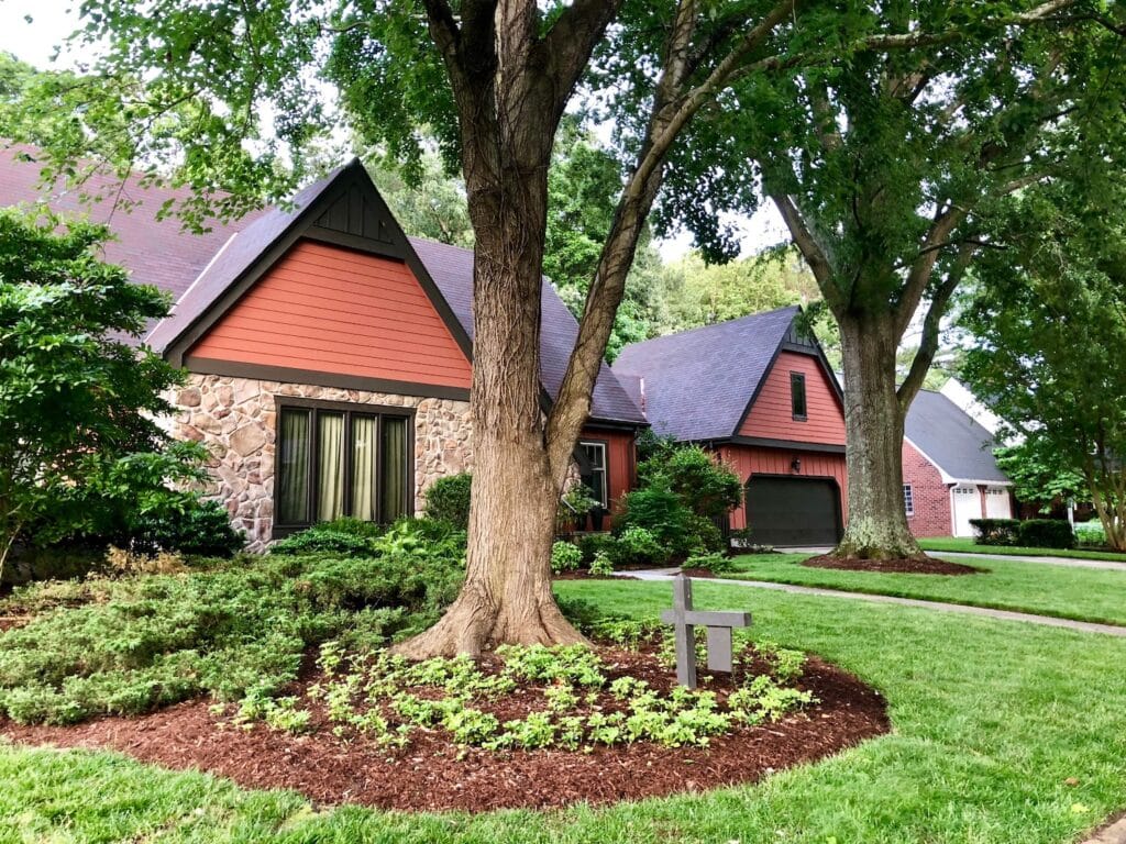 Reddish-orange gabled houses with stone accents sit behind a large mature tree and a lush green lawn in a suburban neighborhood.