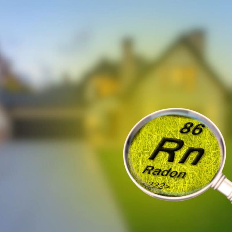 A magnifying glass highlighting the chemical symbol for Radon (Rn) against a blurred residential house background.