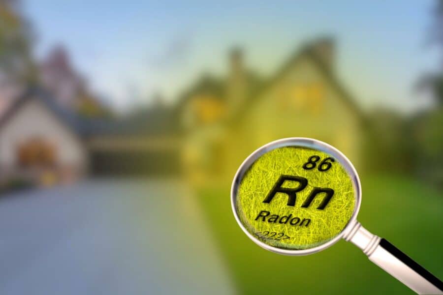 A magnifying glass highlighting the chemical symbol for Radon (Rn) against a blurred residential house background.