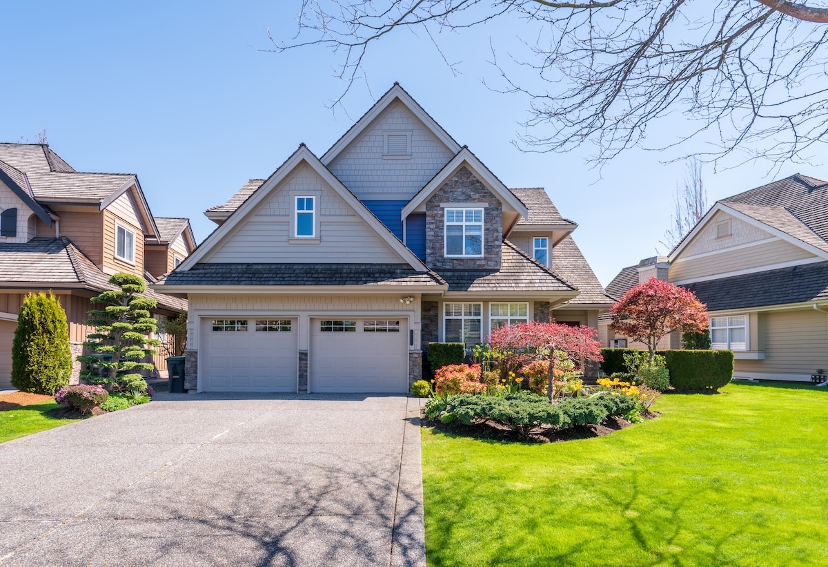 Exterior view of a large, two-story suburban home featuring a mix of gray siding and stone accents, a two-car garage, and a beautifully landscaped front yard on a sunny day.
