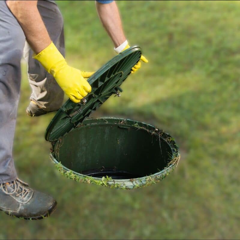 A worker wearing yellow protective gloves lifts the green lid of an underground septic tank in a grassy lawn.