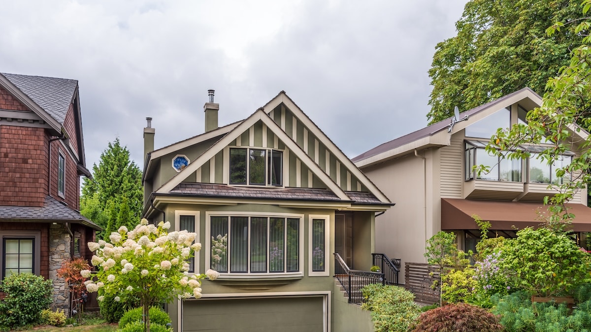 A row of unique multi-story homes, highlighted by a central olive-green house with Craftsman-style architectural trim, a large bay window, and a blooming white hydrangea bush in the front yard.
