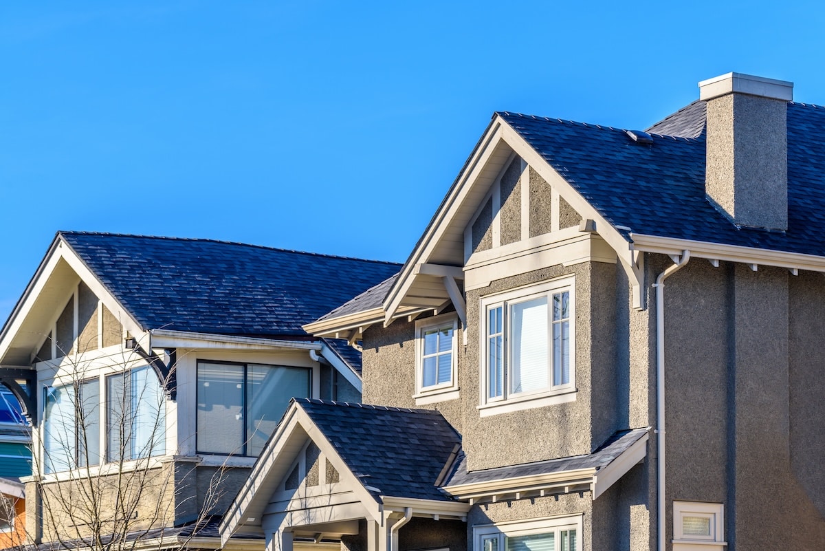 The upper floors and rooflines of modern Craftsman-style homes featuring dark gray architectural shingles, textured gray stucco walls, decorative white gable trim, and a prominent chimney against a bright blue sky.