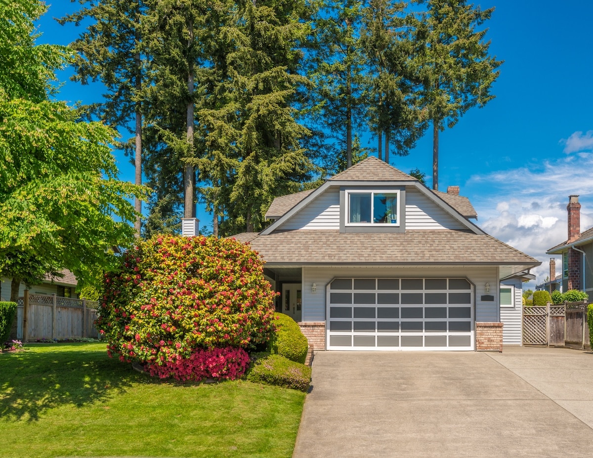 Exterior view of a suburban house featuring a wide concrete driveway, green lawn with vibrant flowering bushes, and tall pine trees under a clear blue sky.