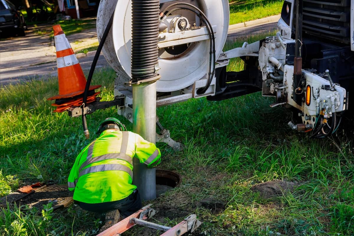 A utility worker wearing a high-visibility jacket guiding a large vacuum hose into an open manhole beside a sewer cleaning truck on a grassy roadside