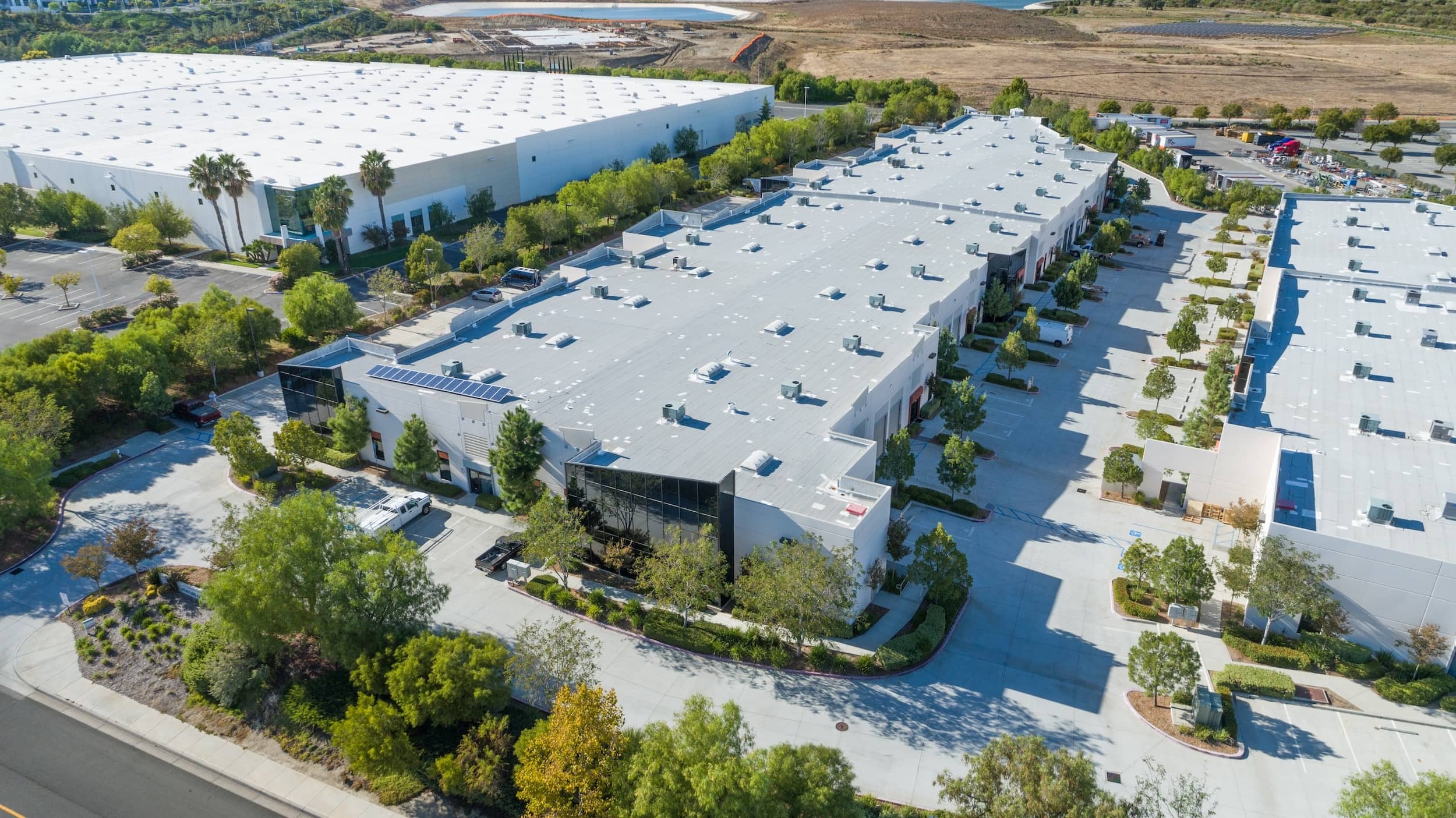 An aerial view of a large industrial business park featuring flat-roofed warehouse buildings, parking lots, and surrounding landscaping.