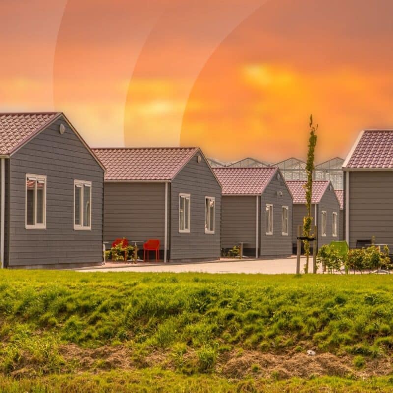 Community of gray modular manufactured homes with dark roofs and green lawn at golden hour