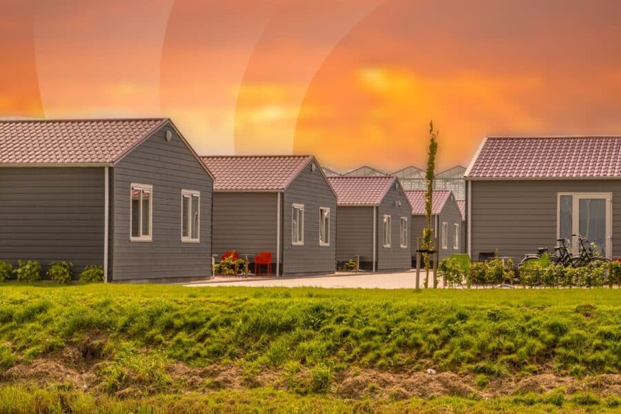 Community of gray modular manufactured homes with dark roofs and green lawn at golden hour