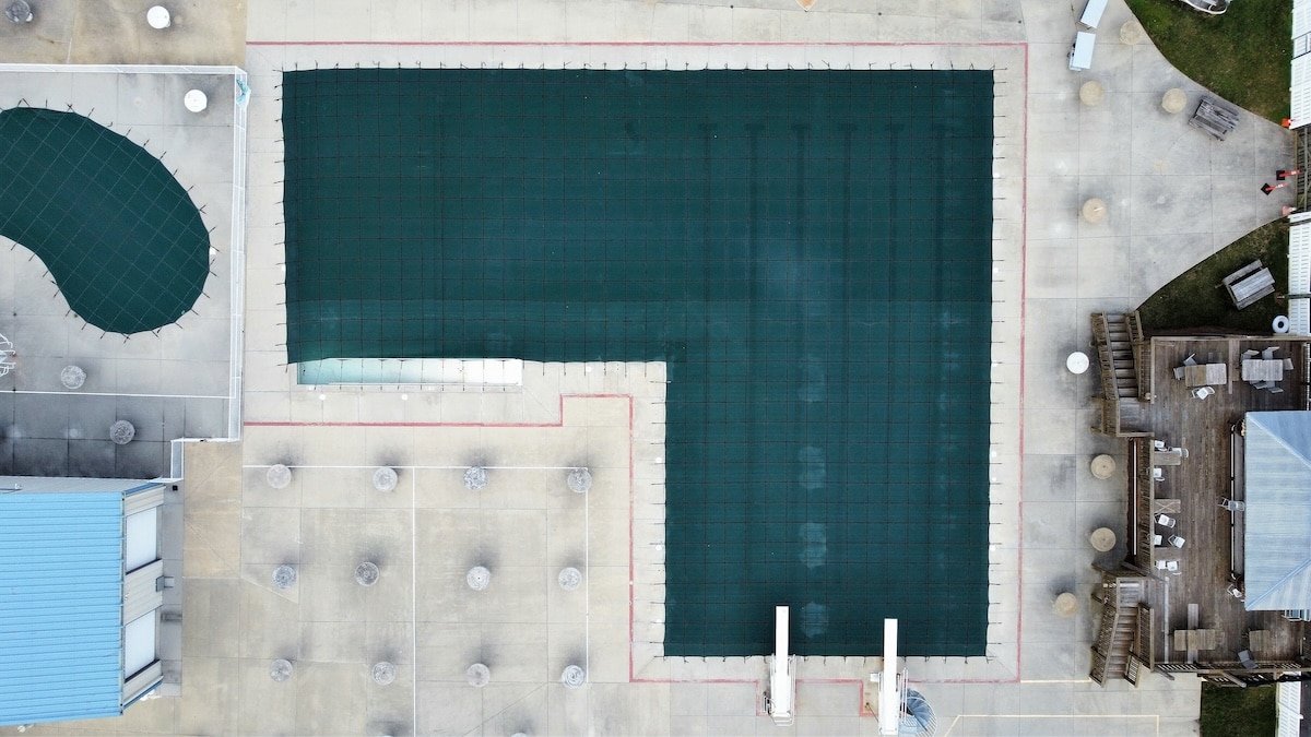 An aerial view of a large L-shaped swimming pool secured with a dark green mesh cover on a concrete deck.