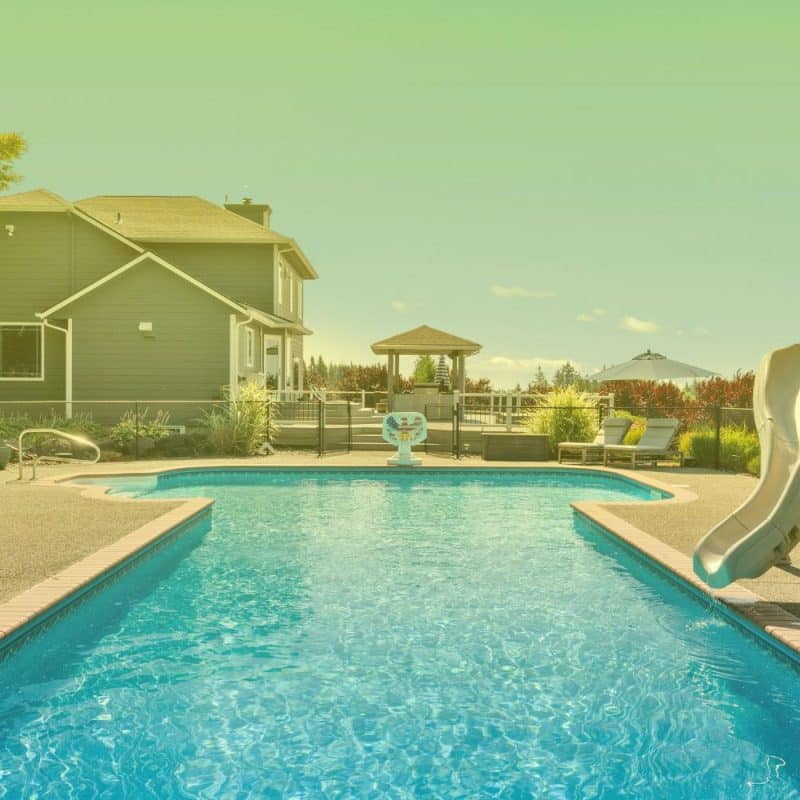 Backyard in-ground swimming pool with clear blue water, a water slide, concrete deck, and a house in the background.