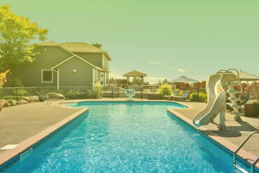 Backyard in-ground swimming pool with clear blue water, a water slide, concrete deck, and a house in the background.